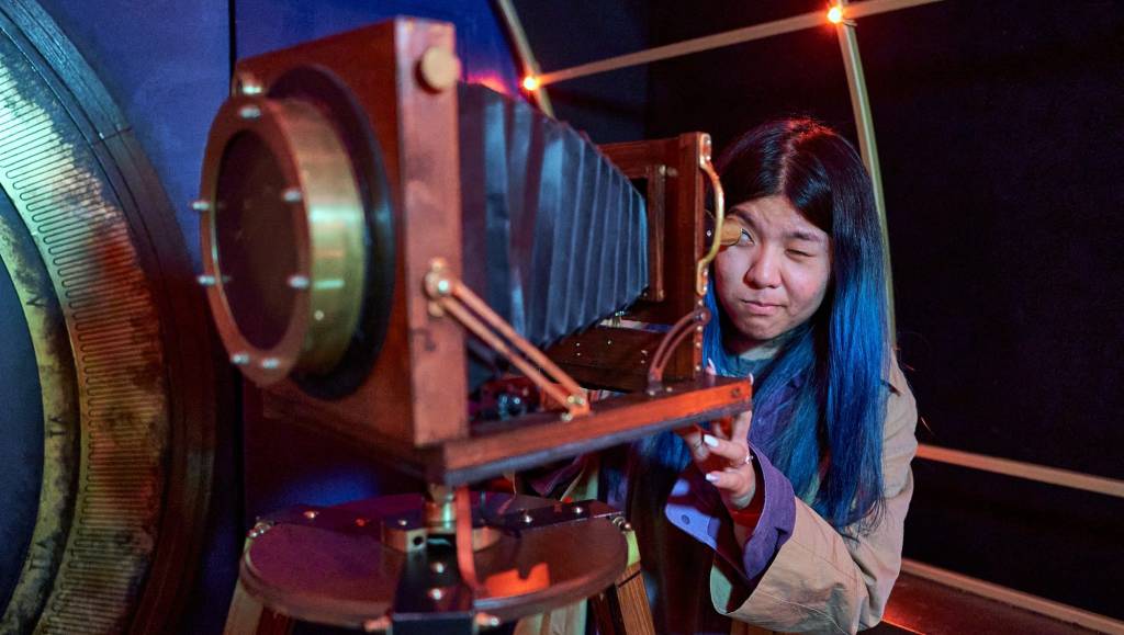 Someone peers through a "telescope" that looks like an old-school camera