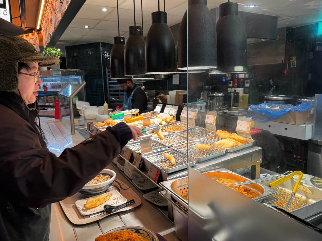 A person picks a tempura prawn off a shelf onto their tray