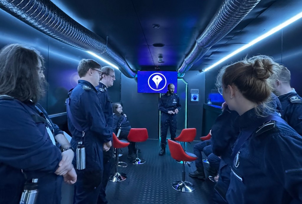 A group of people in a long briefing room, listening to a man at the end standing in front of a screen