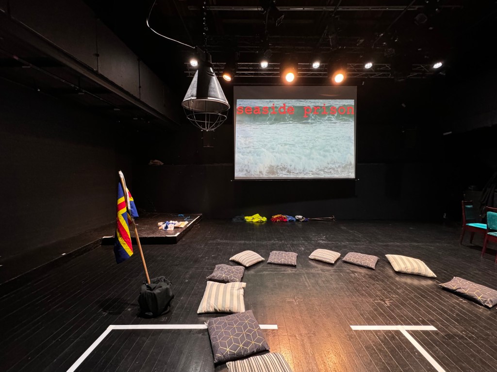 An empty black box theatre with a projector screen on the far wall, a flag, a taped outline of a house, and cushions on the floor in a circle