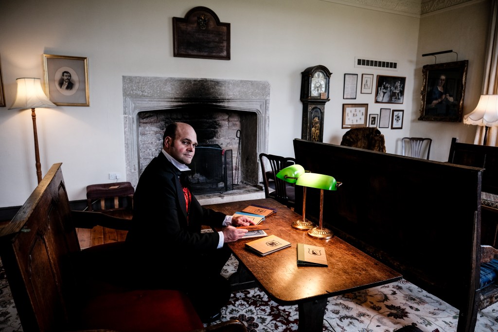 A man in old-style formal clothing sits at a table with a set of books in front of him, in a parlour room. He looks at us enticingly.
