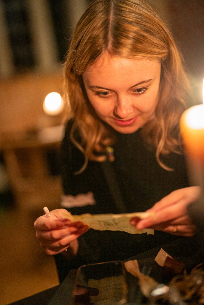 A woman reads a small scroll closely, lit by candles