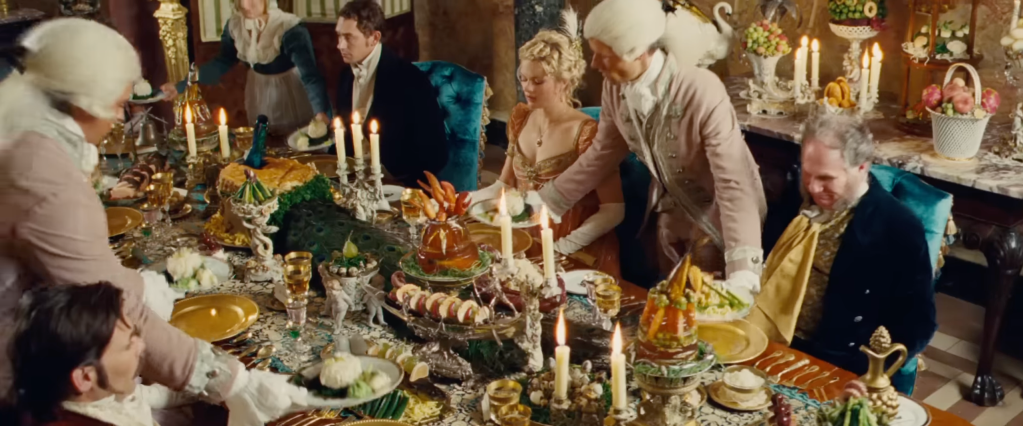 A dining table laden with elaborate dishes. Guests in period costume are sitting down while servants lay down more dishes.