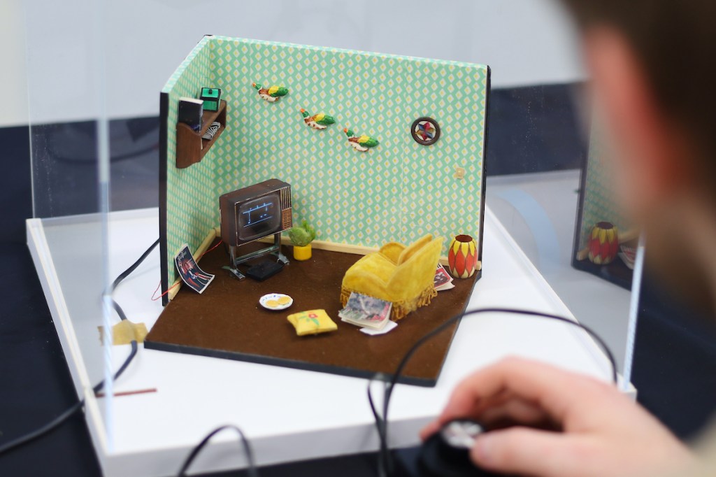 A miniature diorama of a room with a tiny TV, with a person holding a game controller in front of it