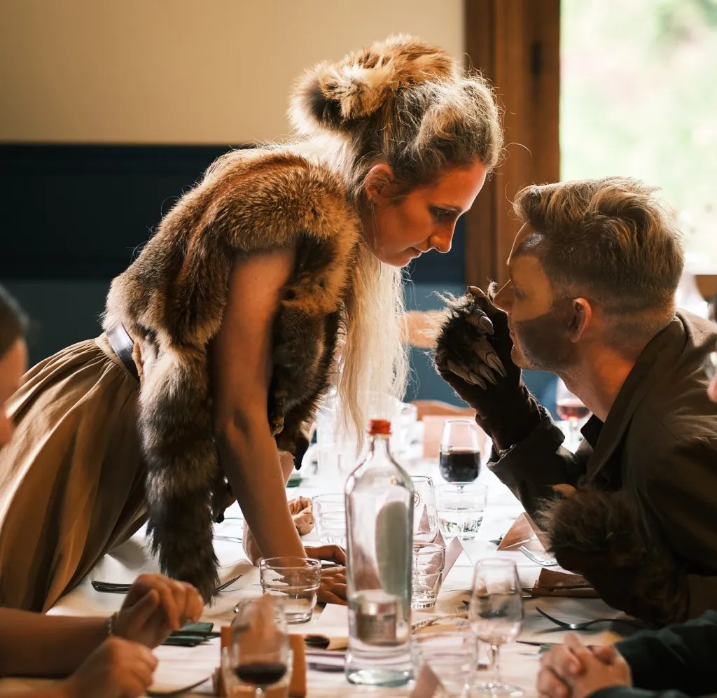 A people in animal costume facing off across a dining table