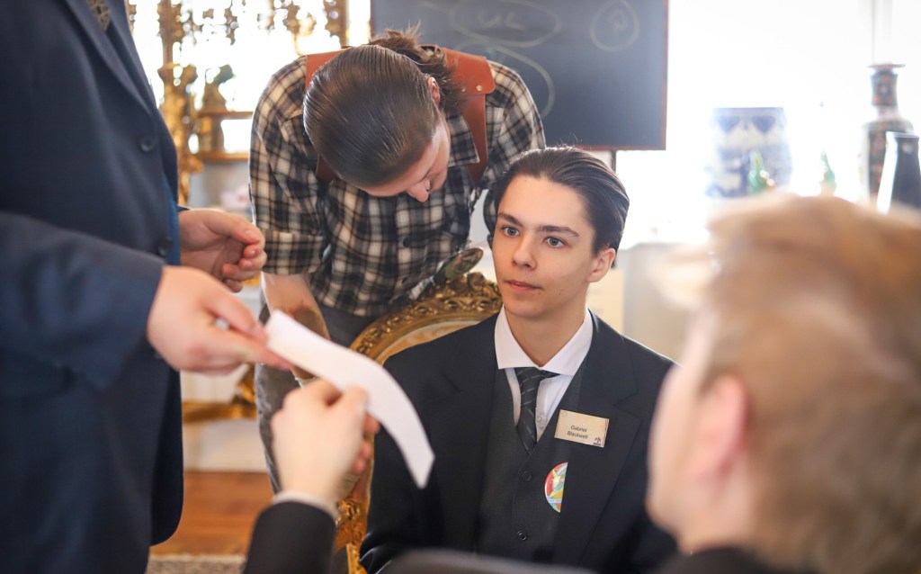 A seated student with tie and jacket and slicked back hair, with another student perching by him looking similar