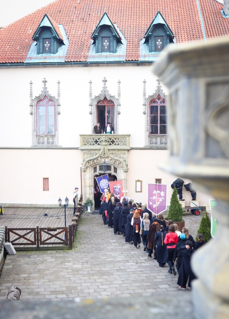 A large old building with students walking to a gate in robes in a line, bearing banners