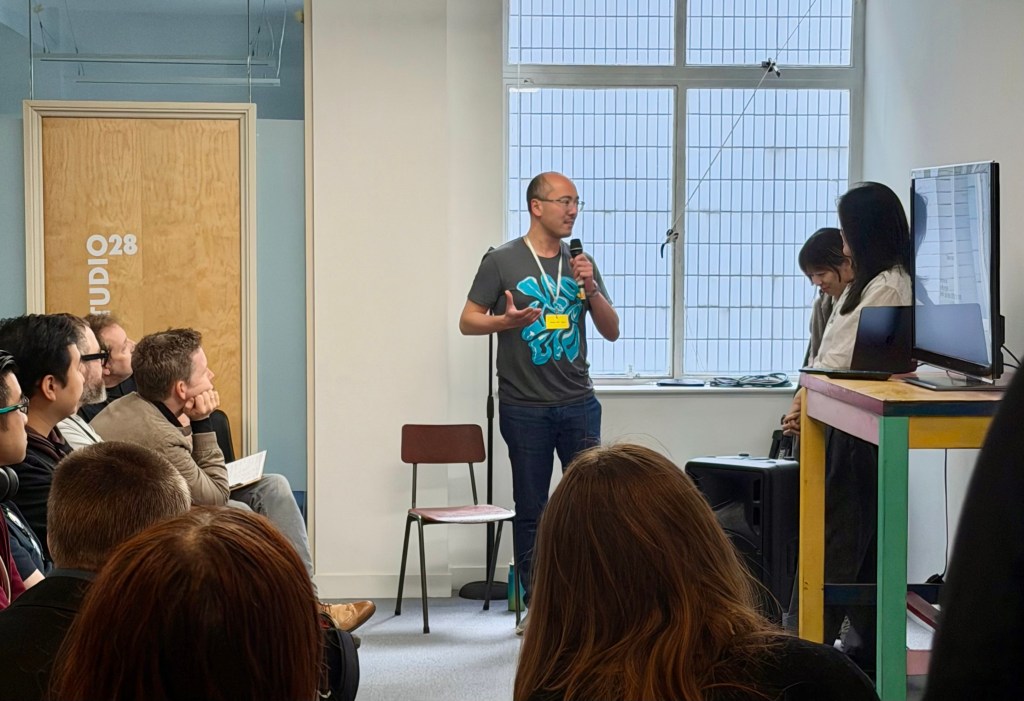 Adrian holding a microphone talking to two speakers in front of an audience