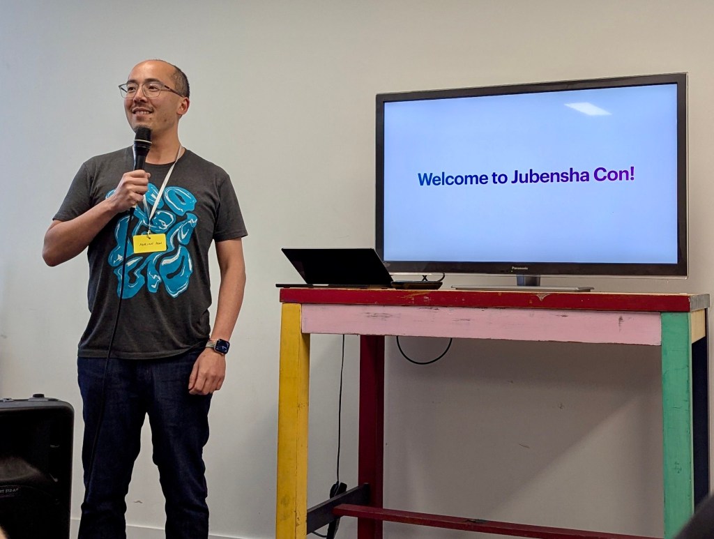 Me holding a microphone next to a TV with the words "Welcome to Jubensha Con!"