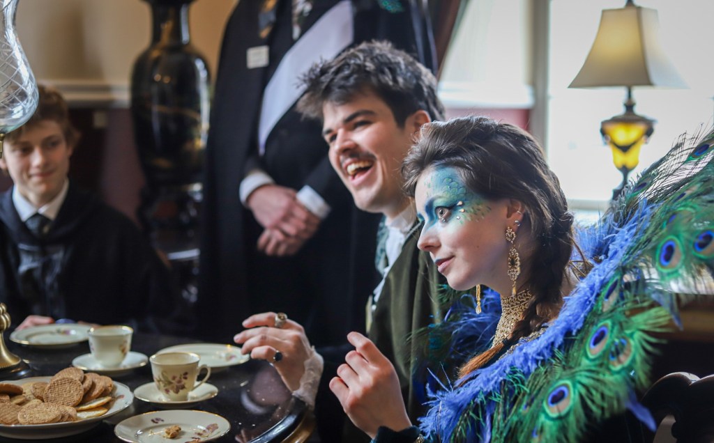 Two people in period costumes, with one person wearing feathers and makeup like a peacock