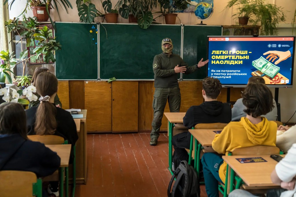 A man in military clothes and a balaclava at the front of a classroom pointing at a TV screen
