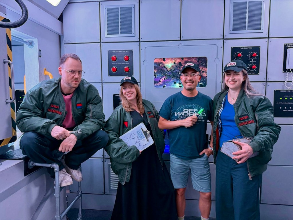 Four people smiling for the camera in space-themed bomber jackets in a spaceship escape room