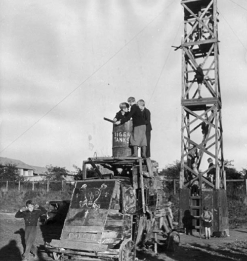 Three children on a shack, and a 50ft tall tower behind them with children climbing through it