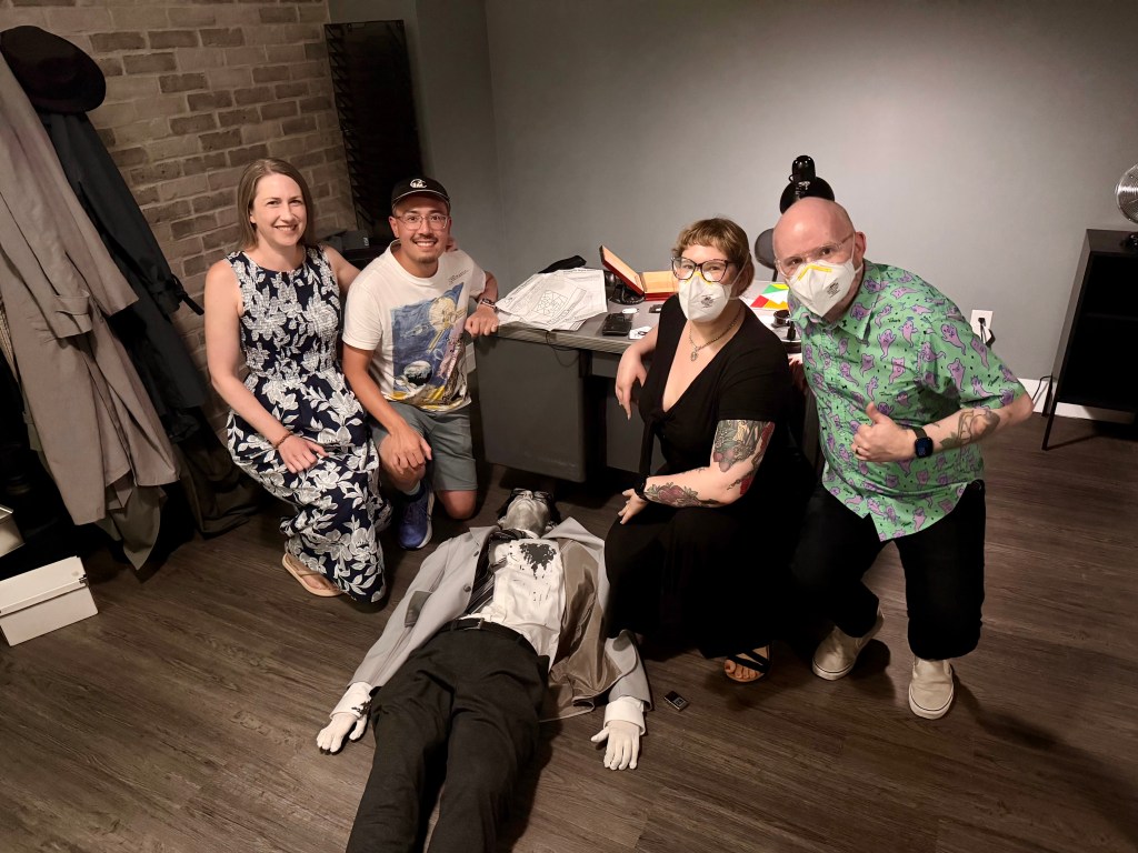 Four people kneeling by a mannequin corpse, smiling at the camera