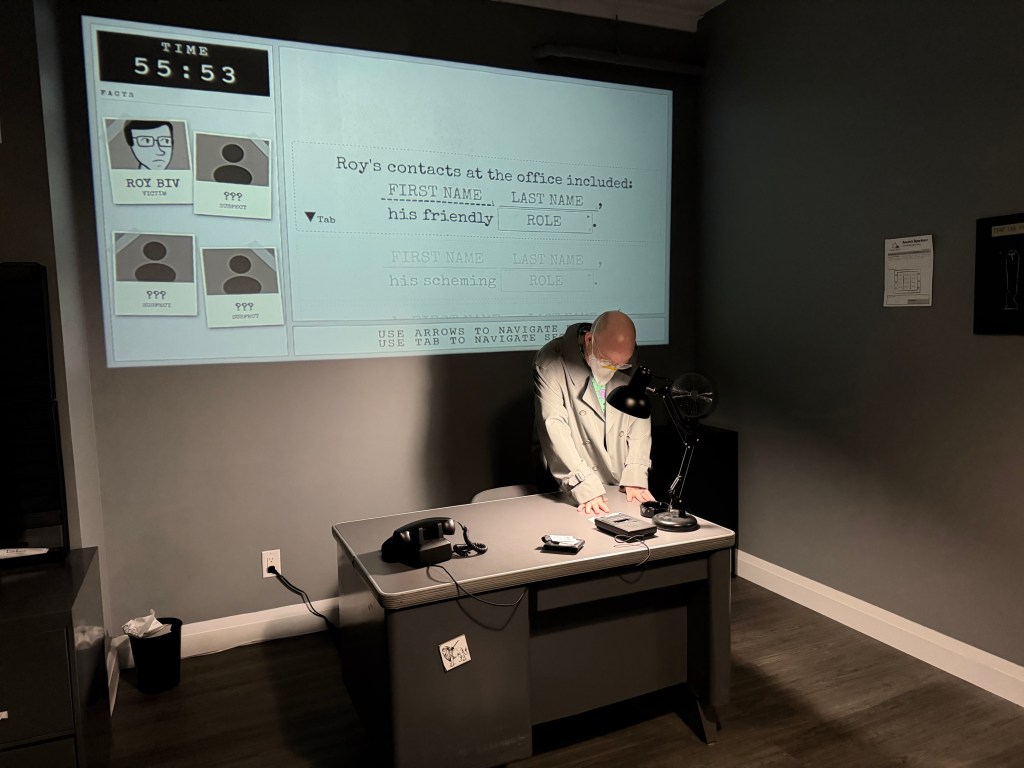 A A black and white-coloured escape room that looks like an office. On the wall is a projected image of a game interface. There are polaroids of characters and names, a countdown, and a mad-libs sentence: "Roy's contacts at the office included FIRST NAME / LAST NAME, his friendly ROLE"