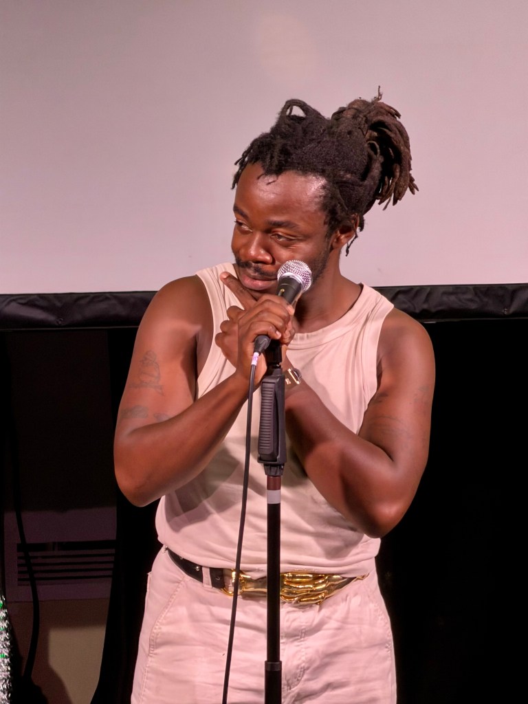 Demi Adejuyigbe in a tanktop with a hand to his chin, looking cheeky