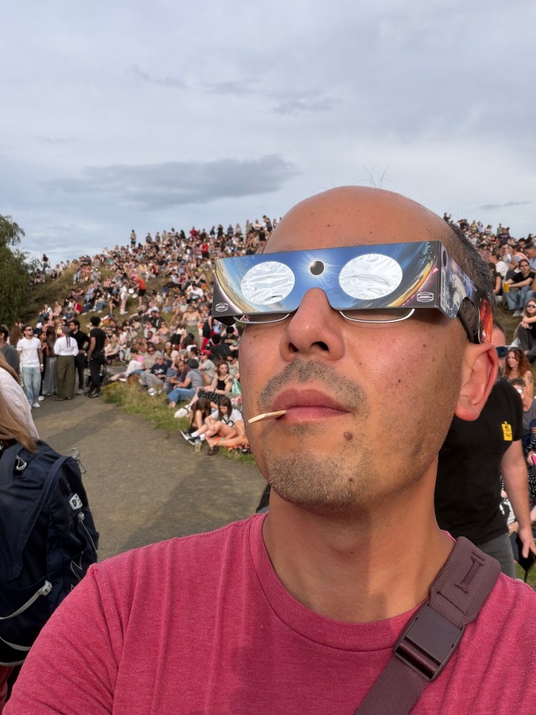 Selfie of me wearing solar eclipse viewing glasses in front of a hill with a crowd sitting on the grass