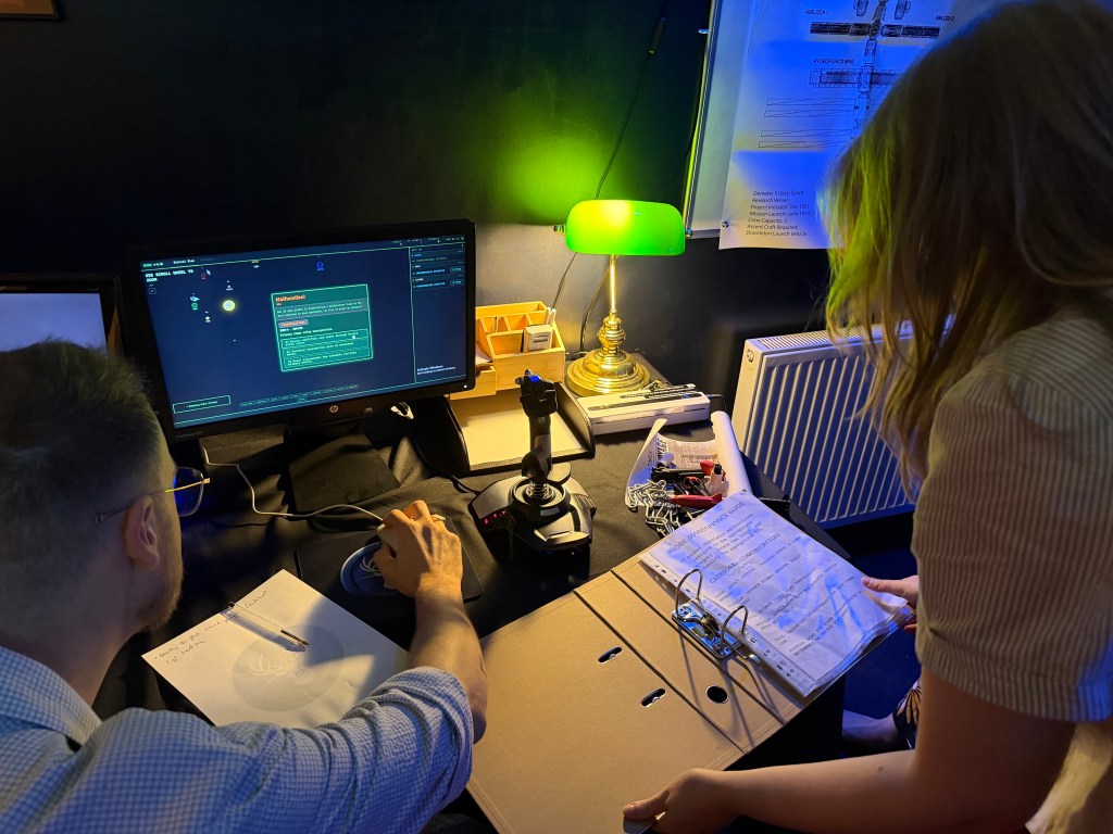 One person controls a computer with a sci-fi interface with the other flips through a set of paper files