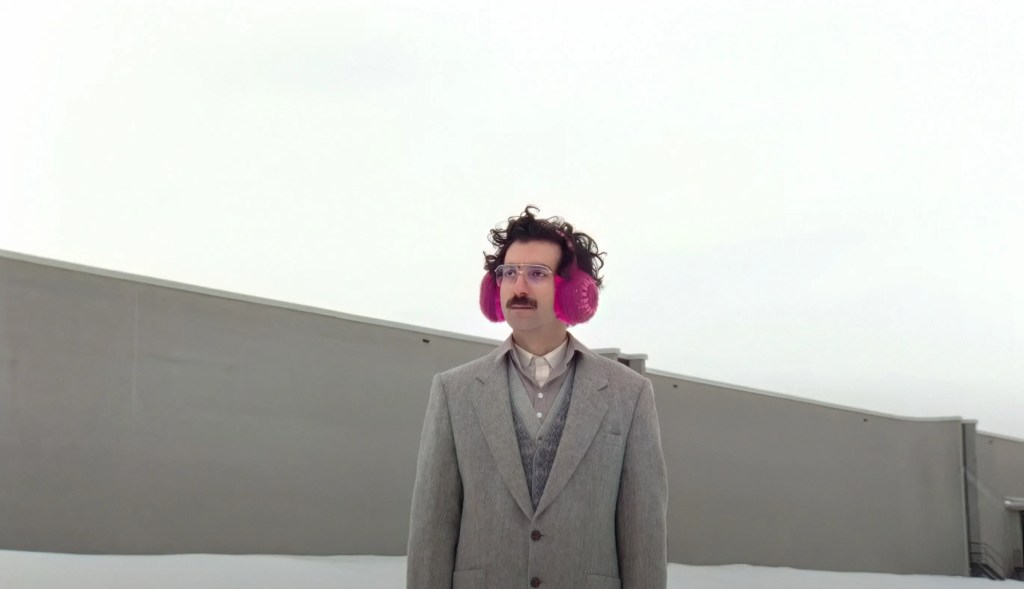 A man in a suit with red earmuffs, standing in front of a grey wall